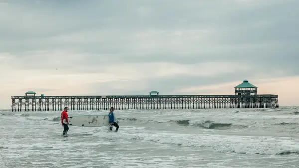 A surfer in the shallows beside the long Folly Beach pier