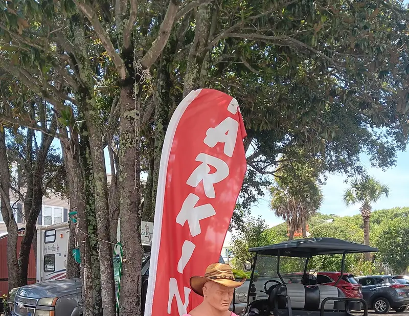 A red feather flag reading PARKING beside hand painted PARK HERE signs under the oaks
